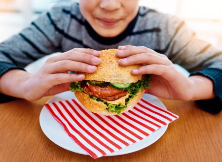 Téléchargez les photos : Enfant, les mains et manger un hamburger sur la table pour un délicieux déjeuner, repas ou nourriture avec des légumes sains à la maison. Main de petit garçon affamé tenant un hamburger de bœuf pour le dîner frais, la nutrition ou les vitamines. - en image libre de droit