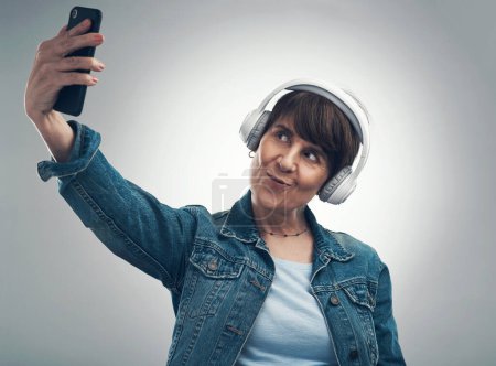 Photo for I know all about the best angles too. Studio shot of a senior woman taking selfies while wearing headphones against a grey background - Royalty Free Image