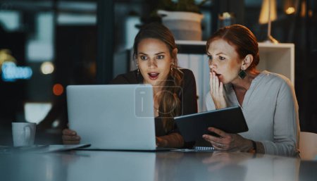 Der Erfolg geht über das 9-5. Zwei Geschäftsfrauen schauen überrascht, während sie nachts gemeinsam an einem Laptop im Büro arbeiten