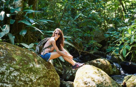 Téléchargez les photos : Prêt pour une aventure. une belle jeune femme assise sur un rocher au bord d'un ruisseau dans la forêt - en image libre de droit
