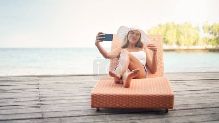 Téléchargez les photos : Jeune femme, selfie lounge et cocktail en vacances au bord de l'océan sur une chaise longue avec un sourire. Femme, voyage et Maldives île en été prendre une photo pour une photo de profil et les médias sociaux. - en image libre de droit