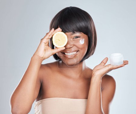 Téléchargez les photos : Les thats hydratants boostés de vitamine c. Plan studio d'une jolie jeune femme tenant un citron et appliquant de l'hydratant sur son visage sur un fond gris - en image libre de droit