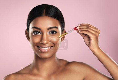 Photo for Face, skincare serum and happy woman in studio isolated on a pink background. Portrait, cosmetics and Indian model with hyaluronic acid, essential oil or vitamin c dropper for healthy dermatology - Royalty Free Image
