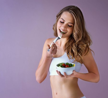 Foto de Come de acuerdo a tus metas. Foto de estudio de una joven saludable comiendo una ensalada sobre un fondo morado - Imagen libre de derechos
