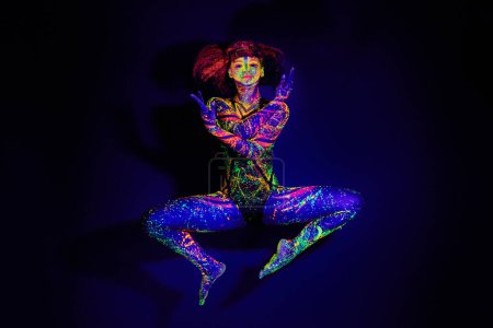 Téléchargez les photos : Peinture corporelle, danse et portrait d'une femme en néon isolé sur un fond sombre dans un studio. Modèle créatif, lumineux et dansant avec art psychédélique pour la créativité et abstrait fluorescent. - en image libre de droit