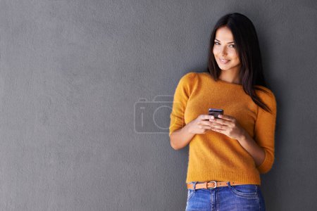 Foto de Es una mariposa social. Una atractiva joven sosteniendo un teléfono móvil mientras está de pie contra una pared gris - Imagen libre de derechos