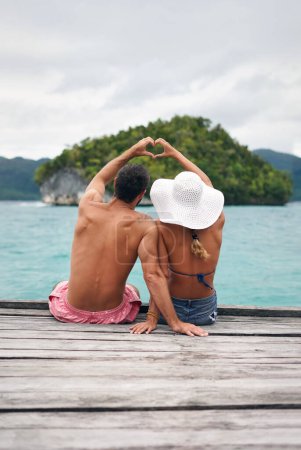 Photo for Heart shape, hands and couple by ocean while on a summer, romantic and tropical vacation. Island, dock and back of man and woman relaxing on wood pier with love gesture on holiday or weekend trip - Royalty Free Image