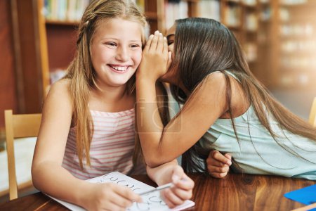Téléchargez les photos : J'ai un secret. une jeune fille joyeuse chuchotant dans l'oreille de ses amis pendant qu'ils sont tous les deux assis à l'intérieur d'une salle de classe pendant la journée - en image libre de droit