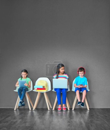 Téléchargez les photos : Grandir avec des gadgets sans fil. Plan studio d'enfants assis sur des chaises et utilisant la technologie sans fil sur un fond gris - en image libre de droit
