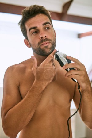 Téléchargez les photos : Couper ma barbe à la perfection. un beau jeune homme se rasant à la maison - en image libre de droit