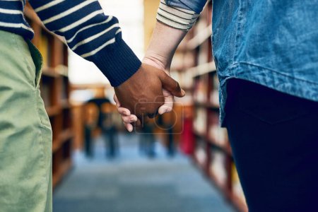 Téléchargez les photos : On étudie mieux quand on était ensemble. un couple se tenant la main alors qu'ils traversent une bibliothèque - en image libre de droit