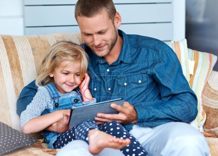 Photo for Two can play this game. an adorable little girl sitting on the sofa using a digital tablet with her father - Royalty Free Image