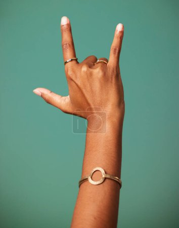 Photo for Rock and roll hand sign by a studio for party, cool or wild greeting with rings and bracelet jewelry. Emoji, punk and woman rebel or rocker gesture with fingers isolated by a dark green background - Royalty Free Image