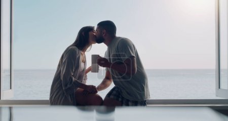 Photo for Couple, kiss and coffee in morning by window with love, romance and care on holiday by ocean with sunshine. Man, woman and trust with drink, espresso or matcha on vacation by sea with bond in summer. - Royalty Free Image