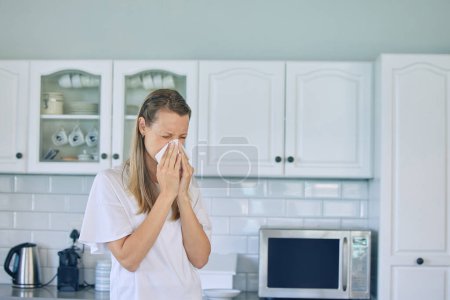 Foto de Alergias, papel higiénico y mujer sonándose la nariz en la cocina por un resfriado, gripe o estornudo en su casa. Enfermedad, virus y mujer joven con tejido para los senos paranasales, fiebre del heno o covidio en el apartamento - Imagen libre de derechos