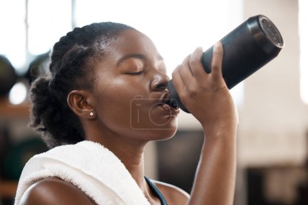 Foto de Mujer negra, acondicionamiento físico y agua potable en el gimnasio para la sostenibilidad, la hidratación o la sed después del ejercicio. Persona africana sedienta con bebida para refrescarse después del entrenamiento cardiovascular en interiores. - Imagen libre de derechos