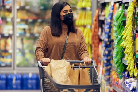 Photo for Grocery shopping, woman and sale at a retail shop, market and store for groceries with mask. Health, virus safety and young female person with cart and food purchase in a supermarket at shelf. - Royalty Free Image