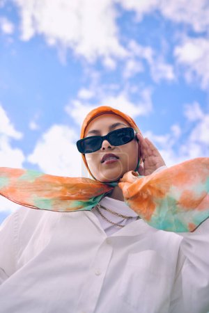 Foto de Retrato, moda o hijab con una mujer saudí al aire libre sobre un fondo de cielo azul en una bufanda y gafas de sol para el estilo. Musulmán, la fe o la religión con una persona árabe joven de moda en el exterior con ropa moderna. - Imagen libre de derechos