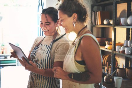 Foto de Vendiendo sus creaciones en el mercado de las redes sociales. dos mujeres jóvenes usando una tableta digital mientras trabajan juntas en un estudio de cerámica - Imagen libre de derechos
