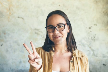 Photo for The happy-go-lucky have the best luck. Portrait of a young woman making a peace sign while standing against a wall - Royalty Free Image