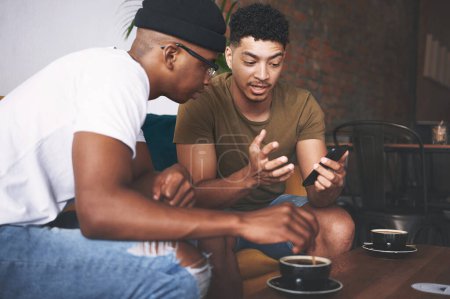 Foto de Tiene muchas características geniales. hombres discutiendo algo en un celular mientras están sentados juntos en una cafetería - Imagen libre de derechos