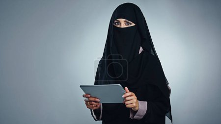 Téléchargez les photos : Vivre en ligne avec les dernières technologies. Portrait studio d'une jeune femme portant une burqa et utilisant une tablette numérique sur fond gris - en image libre de droit