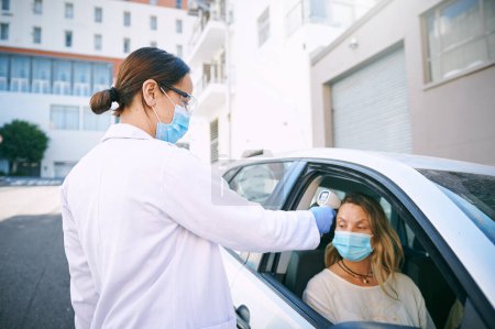 Téléchargez les photos : Avant d'entrer, il faut en être sûr. une jeune femme masquée se faisant contrôler sa température par un médecin assis dans sa voiture - en image libre de droit