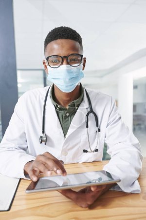 Photo for Shall we go over your test results. a young doctor wearing a face mask and sitting in his office at the clinic while using technology - Royalty Free Image