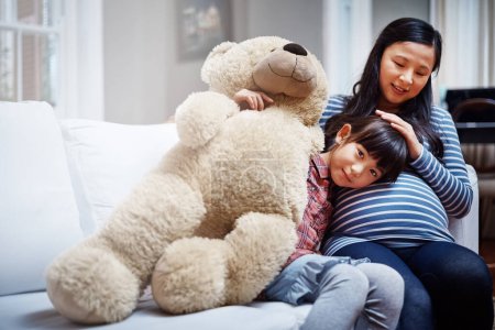 Photo for Theres a brother or sister in there for me. Cropped portrait of a little girl sitting with her mother and her teddybear on the sofa - Royalty Free Image