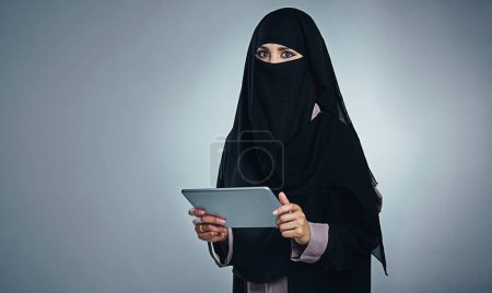 Téléchargez les photos : Lié à tout ce qui est courant. Portrait studio d'une jeune femme portant une burqa et utilisant une tablette numérique sur fond gris - en image libre de droit