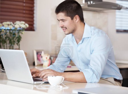 Téléchargez les photos : Profiter des avantages d'Internet. Un beau mâle assis dans sa cuisine avec une tasse de café parcourant sur son ordinateur portable - en image libre de droit