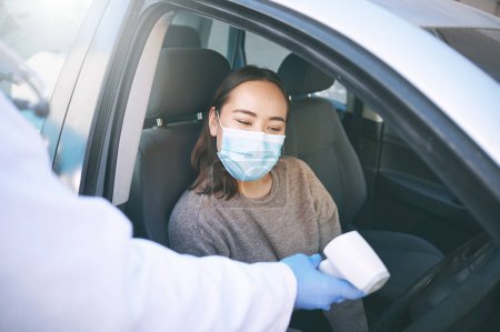 Téléchargez les photos : Restez vigilants, restez en sécurité. une jeune femme masquée se faisant contrôler sa température par un médecin assis dans sa voiture - en image libre de droit