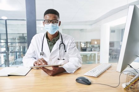 Photo for My job is to save lives. a young doctor wearing a face mask and sitting in his office at the clinic while using technology - Royalty Free Image
