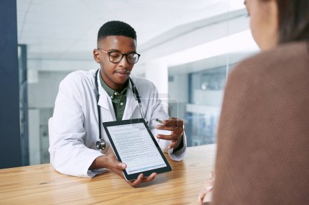 Foto de Por favor, rellene esta lista de verificación Covid. un joven médico sentado con su paciente y usando una tableta digital durante una consulta en la clínica - Imagen libre de derechos
