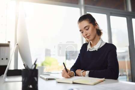Foto de Comprobando sus notas. una atractiva joven empresaria sentada en su escritorio y escribiendo notas en una oficina moderna - Imagen libre de derechos