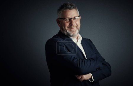 Photo for The corporate world has been good to me. Studio portrait of a handsome mature businessman standing with his arms folded against a dark background - Royalty Free Image