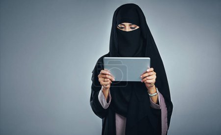 Téléchargez les photos : Connectez-vous et mettez-vous à jour. Plan studio d'une jeune femme portant une burqa et utilisant une tablette numérique sur un fond gris - en image libre de droit