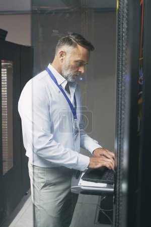 Téléchargez les photos : Sécuriser une connexion stable. un homme mûr utilisant un ordinateur portable tout en travaillant dans une salle de serveurs - en image libre de droit