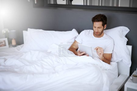 Foto de Navegando a través de sus noticias. un hombre guapo de mediana edad usando su tableta digital mientras se relaja en su dormitorio - Imagen libre de derechos