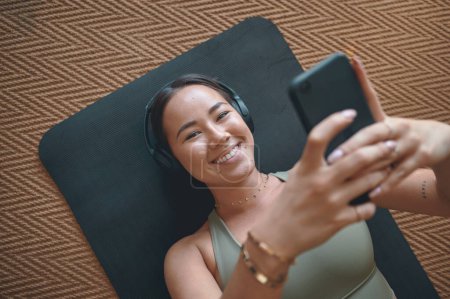 Photo for The right music makes all the difference to your fitness game. Portrait of a young woman wearing headphones and using a cellphone while lying on an exercise mat at home - Royalty Free Image