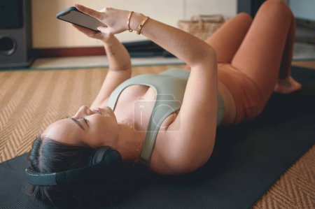 Foto de Desplazamiento a través de aplicaciones de fitness en abundancia. una mujer joven que usa auriculares y un teléfono celular mientras está acostada en una alfombra de ejercicio en casa - Imagen libre de derechos