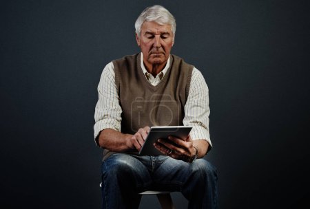 Photo for Hes in touch with the times. Studio shot of a handsome mature man using a tablet while sitting on a wooden stool against a dark background - Royalty Free Image