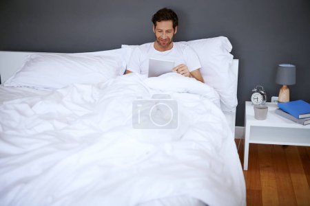 Foto de Este es un buen libro electrónico. un hombre guapo de mediana edad usando su tableta digital mientras se relaja en su dormitorio - Imagen libre de derechos