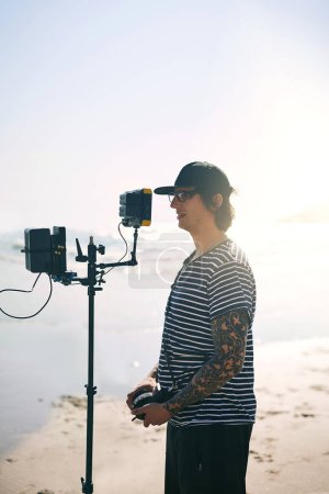 Téléchargez les photos : En attendant le plan parfait. un jeune homme concentré filmant une scène avec une caméra vidéo de pointe à l'extérieur sur une plage pendant la journée - en image libre de droit
