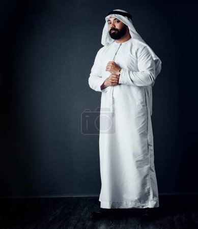 Téléchargez les photos : Il porte bien ses vêtements traditionnels. Plan studio d'un jeune homme vêtu de vêtements traditionnels islamiques posant sur un fond sombre - en image libre de droit