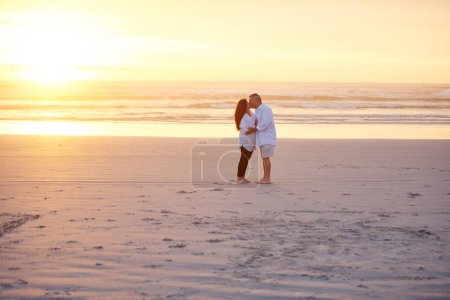 Téléchargez les photos : Rien ne dit plus romantique qu'un baiser au coucher du soleil. un couple mature s'embrassant sur la plage - en image libre de droit