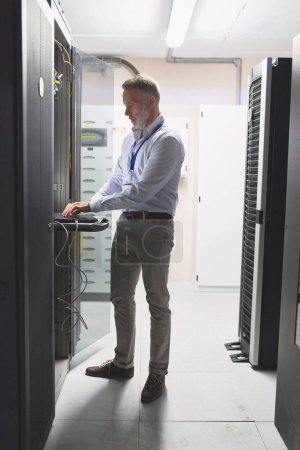 Photo for Processing a system check. a mature man using a laptop while working in a server room - Royalty Free Image