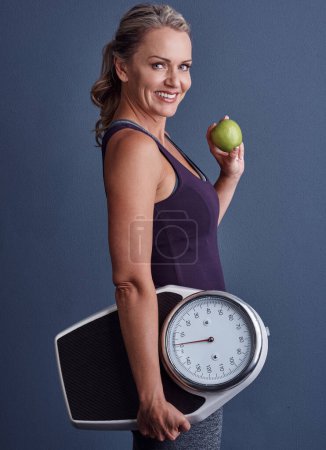 Foto de Dale a tu cuerpo las cosas buenas. Retrato de estudio de una atractiva mujer madura sosteniendo una manzana y una báscula de peso sobre un fondo azul - Imagen libre de derechos
