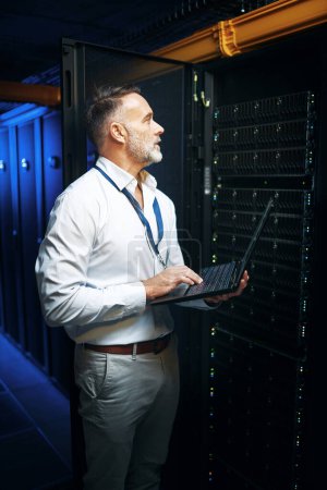 Foto de Hay una cierta manera de mantener todo conectado. un hombre maduro usando un ordenador portátil mientras trabaja en una sala de servidores - Imagen libre de derechos