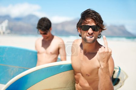 Foto de Playa, dedo medio y hombre surfeando amigos al aire libre juntos para vacaciones de verano o viaje de vacaciones en el extranjero. Surf, mar o grosero con un joven surfista masculino en gafas de sol y amigo vinculación en la costa. - Imagen libre de derechos